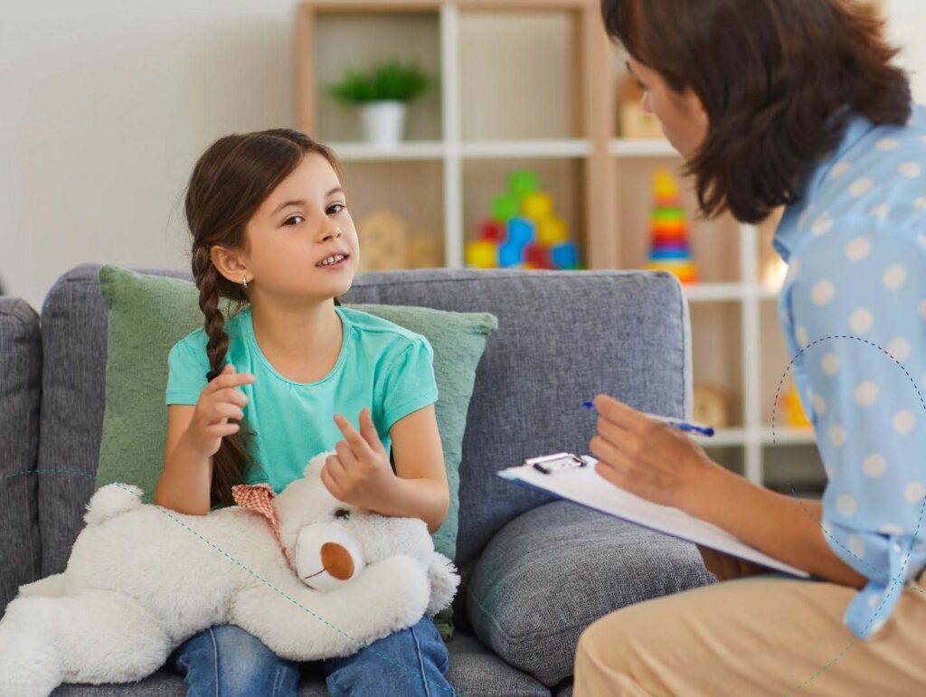 A child sitting on a couch, holding a stuffed bear, talking with a child behavioral health analyst.